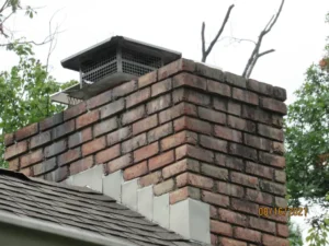 Old brick chimney in disrepair with metal cap and surrounding roof shingles.
