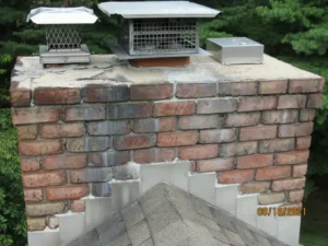 Old brick chimney with signs of disrepair, featuring metal chimney caps and surrounded by trees.
