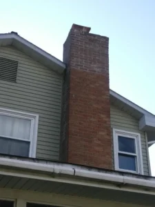 Old, weathered brick chimney on a house with light green siding.