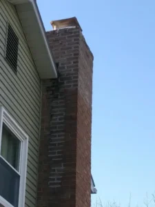 An old, weathered chimney protruding from a building with gray siding under a clear blue sky.