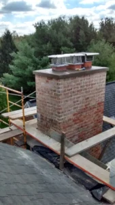 Brick chimney with metal caps and scaffolding on a rooftop with evergreen trees in the background.