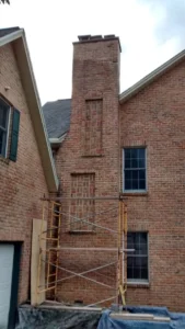 A weathered brick chimney in disrepair with scaffolding for repair against a brick house.