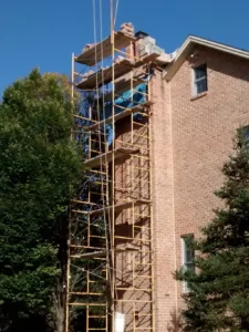 Brick house chimney with scaffolding for reconstruction work, surrounded by a green lawn and bushes.