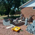 Two workers are building a block wall beside a brick house, surrounded by tools and construction materials in a sunny setting.