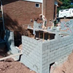 Three workers build a concrete block structure in a backyard, with scaffolding and tools visible under a clear blue sky.