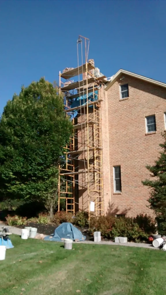 Brick house chimney with scaffolding for reconstruction work, surrounded by a green lawn and bushes.